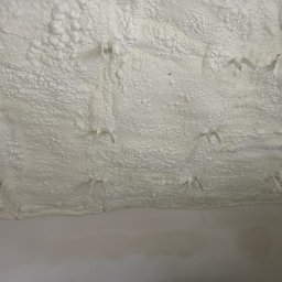 Purisol Karczewski Patryk - Poddasze z ociepleniem pianką PUR, widoczne wystające elementy montażowe. Biała, porowata struktura pianki pokrywa całą powierzchnię. Fragment głowy osoby w prawym dolnym rogu.