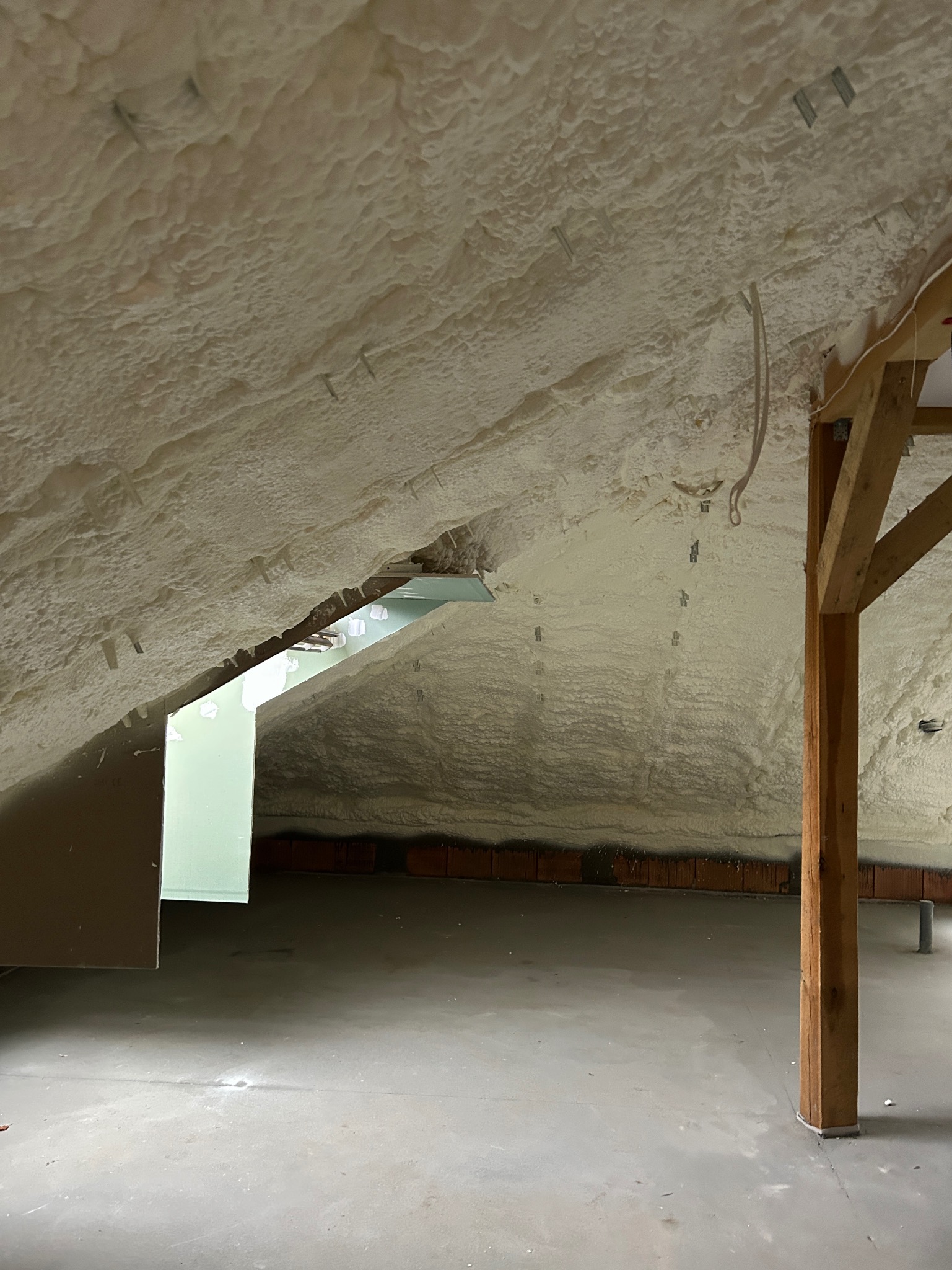 Poddasze po natrysku pianki izolacyjnej, widoczne elementy konstrukcji drewnianej, fragment okna dachowego i ślady po mocowaniach na ścianach.