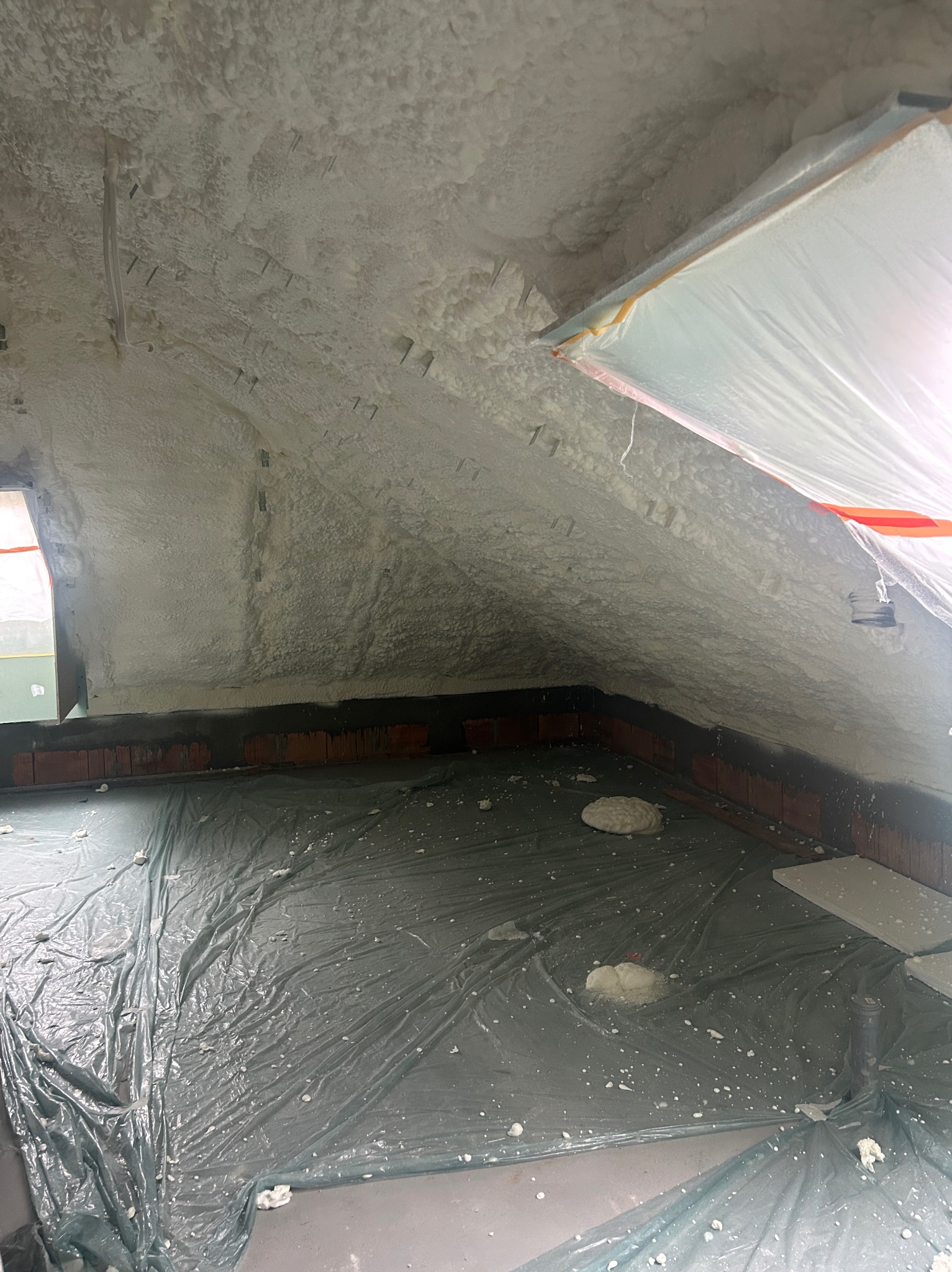 Poddasze po ociepleniu pianką PUR, widoczne rozpylone pozostałości pianki na folii ochronnej podłogi oraz wystające metalowe elementy montażowe w pianie na skośnym suficie.