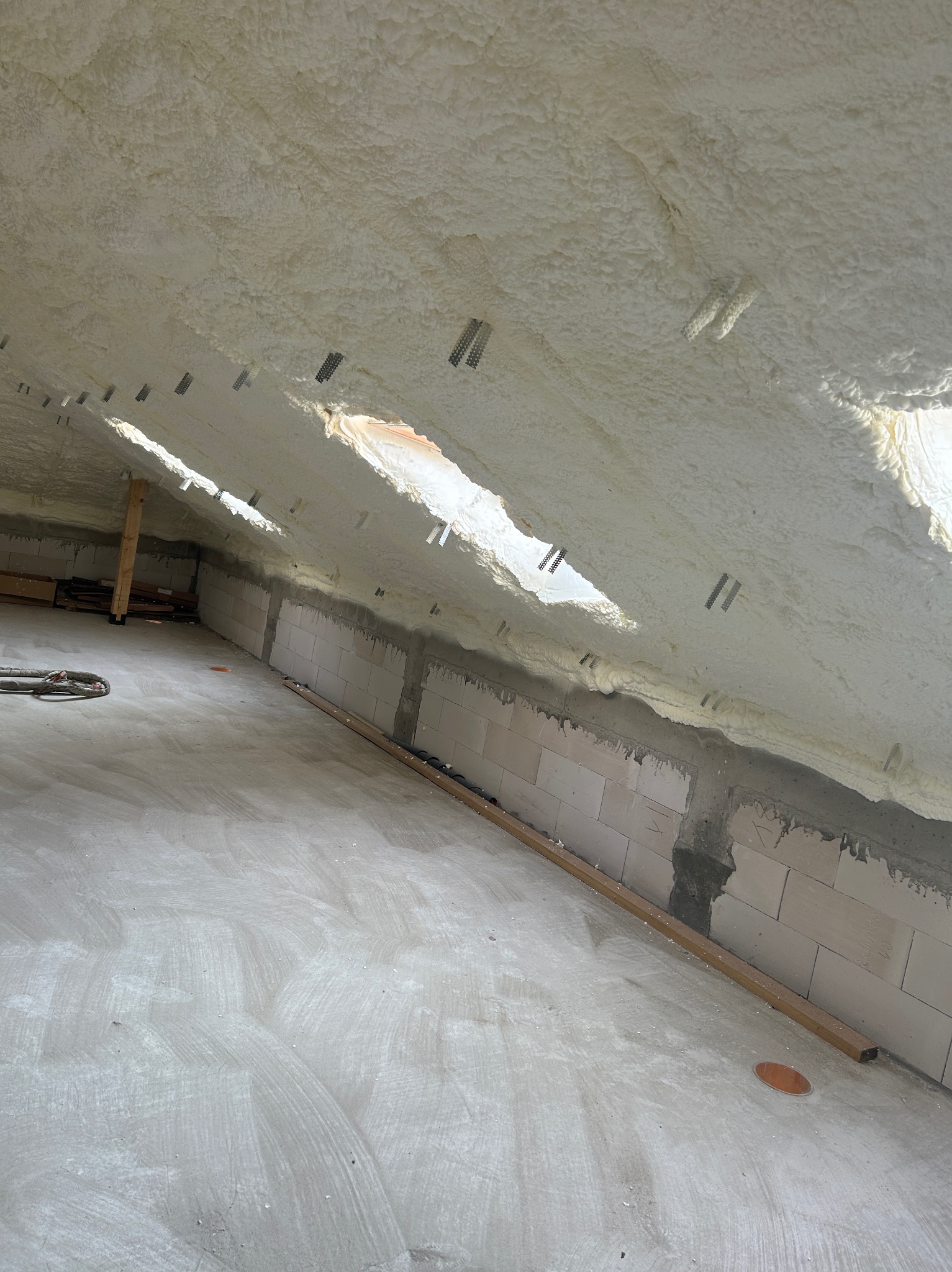 Poddasze po ociepleniu pianką PUR, widoczne okna dachowe i elementy konstrukcyjne. Detal: metalowe mocowania w piance. Świeżo wylana posadzka.