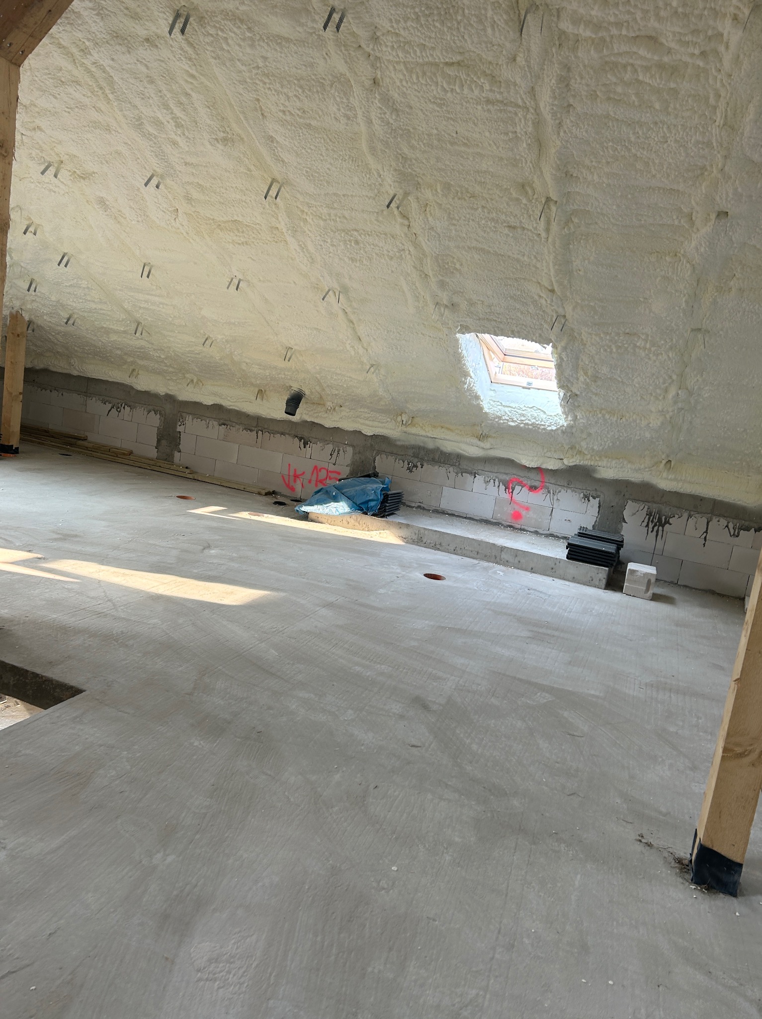 Poddasze po ociepleniu pianką PUR, widoczne krokwie i okno dachowe. Ściany z bloczków betonowych częściowo zakryte izolacją. Betonowa podłoga w surowym stanie.