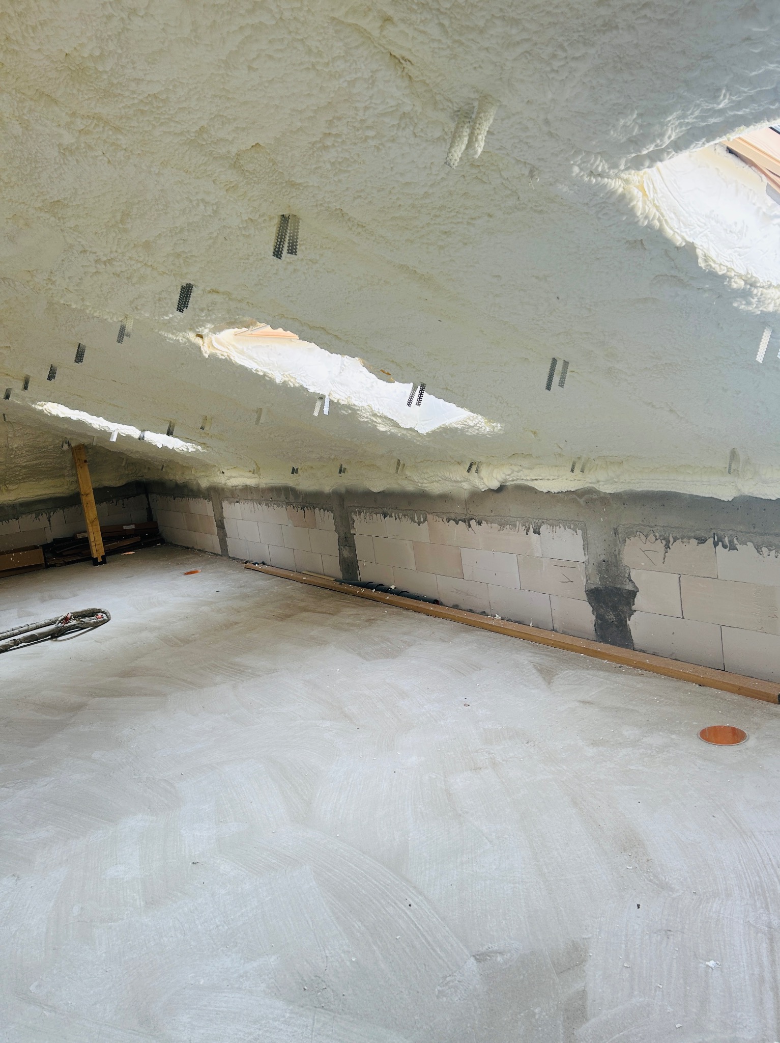 Poddasze po ociepleniu pianką PUR, widoczne krokwie i okno dachowe. Biała pianka pokrywa skośny sufit, ściany z bloczków częściowo zakryte pianką.