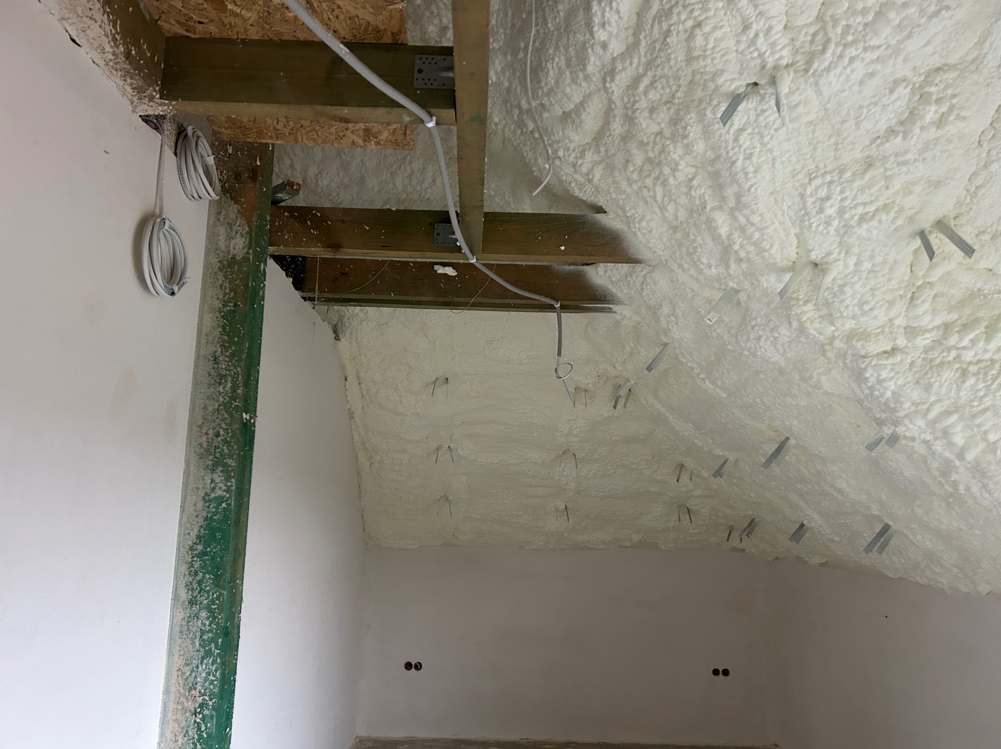 Poddasze po ociepleniu pianką PUR, widoczne drewniane belki stropowe i kable elektryczne. Metalowe elementy mocujące piankę. Białe ściany w surowym stanie.