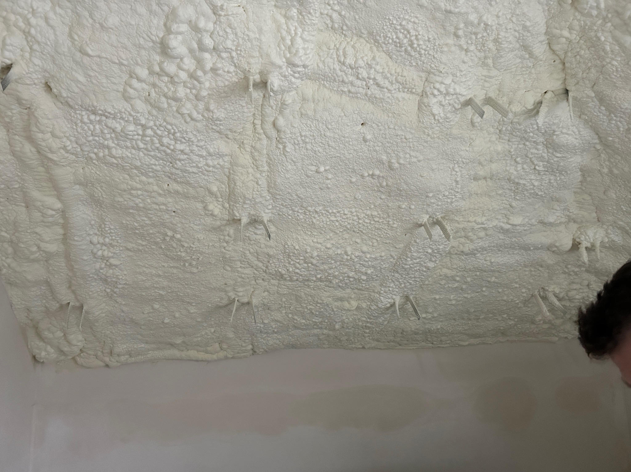 Poddasze z ociepleniem pianką PUR, widoczne wystające elementy montażowe. Biała, porowata struktura pianki pokrywa całą powierzchnię. Fragment głowy osoby w prawym dolnym rogu.