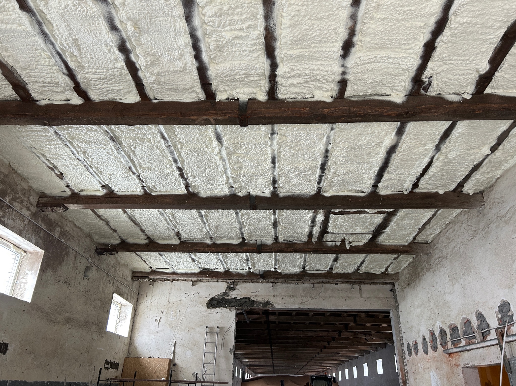 Poddasze budynku gospodarczego z drewnianą konstrukcją, ocieplone białą pianką PUR. Widoczne fragmenty surowych ścian i okna. Realizacja ocieplenia w starym obiekcie.