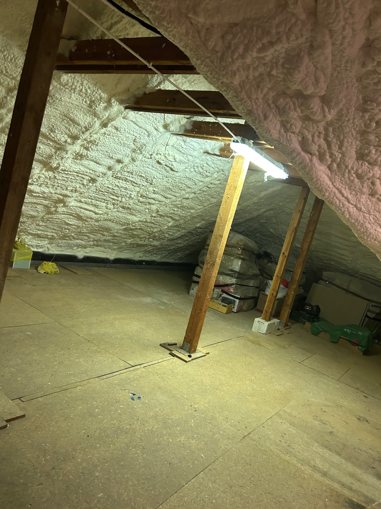 Poddasze ocieplone pianką PUR, widoczne drewniane elementy konstrukcyjne i podłoga z płyt. Oświetlenie sztuczne, w tle materiały budowlane.