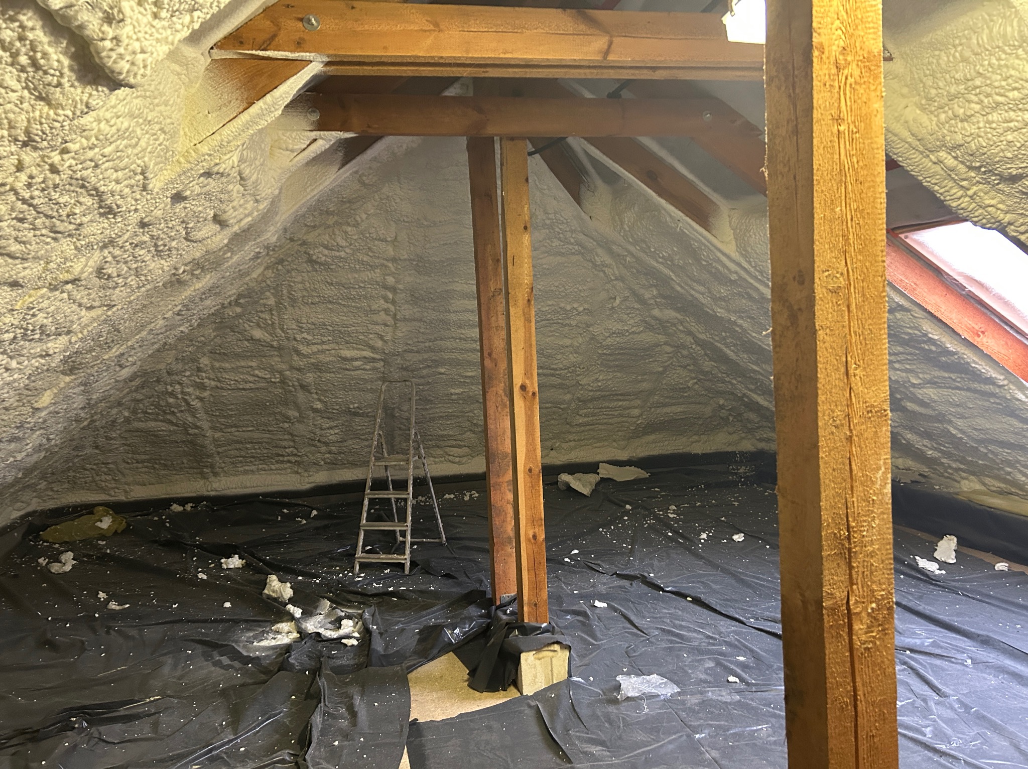 Poddasze po ociepleniu pianką PUR, widoczne drewniane elementy konstrukcyjne i czarna folia ochronna na podłodze. Metalowa drabina oparta o ścianę.