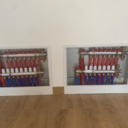 A.T.M.O. Technika Grzewcza - Dwa kolektory ogrzewania podłogowego w białych szafkach, z czerwonymi i niebieskimi rurami, zamontowane na ścianie nad drewnopodobną podłogą.