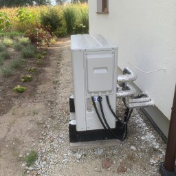 A.T.M.O. Technika Grzewcza - Biała pompa ciepła YORK zamontowana przy ścianie domu, z widocznymi rurami w izolacji. W tle ogród z roślinami i kwiatami. Ujęcie z poziomu gruntu.