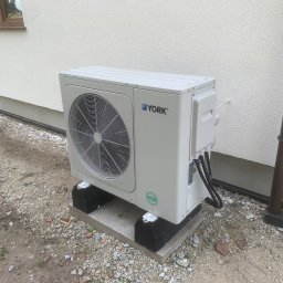 A.T.M.O. Technika Grzewcza - Biała pompa ciepła York R32, zamontowana na betonowej podstawie z czarnymi podkładkami, stoi przy białej ścianie budynku z brązową rurą spustową.