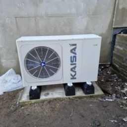 Zewnętrzna jednostka pompy ciepła Kaisai zamontowana na betonowej podstawie z czarnymi podkładkami antywibracyjnymi, przy szarej ścianie budynku w trakcie budowy.