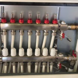 Rozdzielacz ogrzewania podłogowego w szafce instalacyjnej, widoczne zawory, rury i przepływomierze z czerwonymi wskaźnikami.