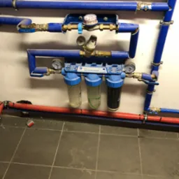 Instalacja filtrów wody z manometrami, rurami w niebieskiej izolacji i zaworami, widoczne różnice w stopniu zabrudzenia wkładów filtracyjnych.