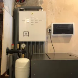 Szara skrzynka sterownicza z logo firmy i manometrem, rury instalacyjne w ścianie, zbiornik wyr&oacute;wnawczy i czarna skrzynka elektryczna na tle ściany w trakcie remontu.