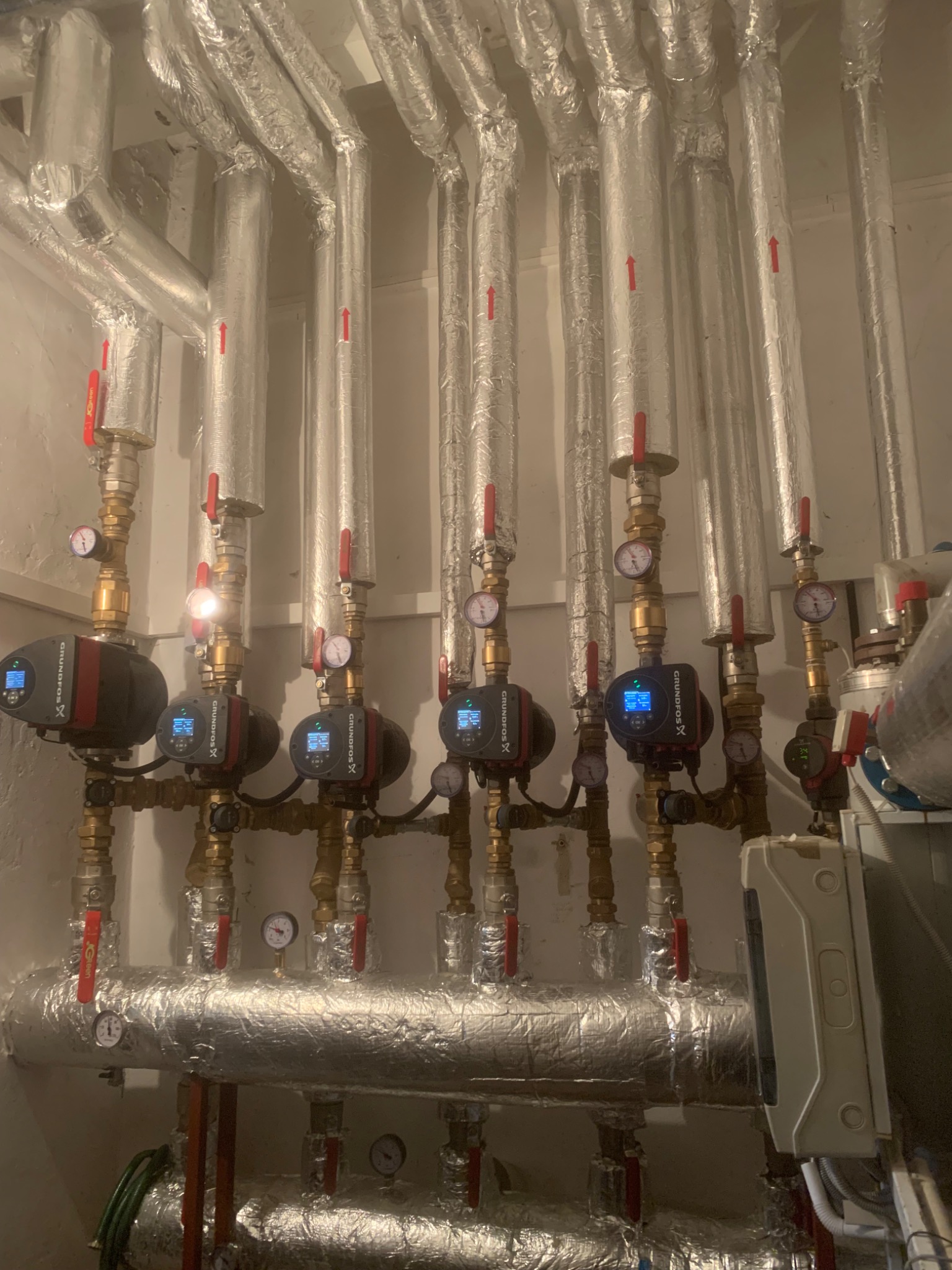 Złożona instalacja grzewcza z pompami obiegowymi Grundfos, rurami w izolacji aluminiowej, zaworami z czerwonymi uchwytami i manometrami w kotłowni.