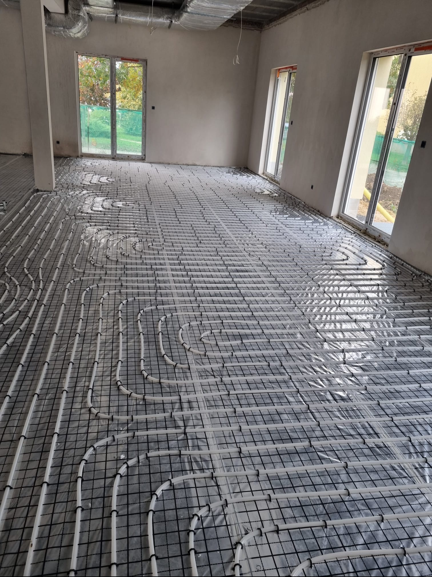 Białe rury ogrzewania podłogowego ułożone na czarnej siatce w surowym wnętrzu z oknami. Widoczne elementy instalacji pod sufitem.