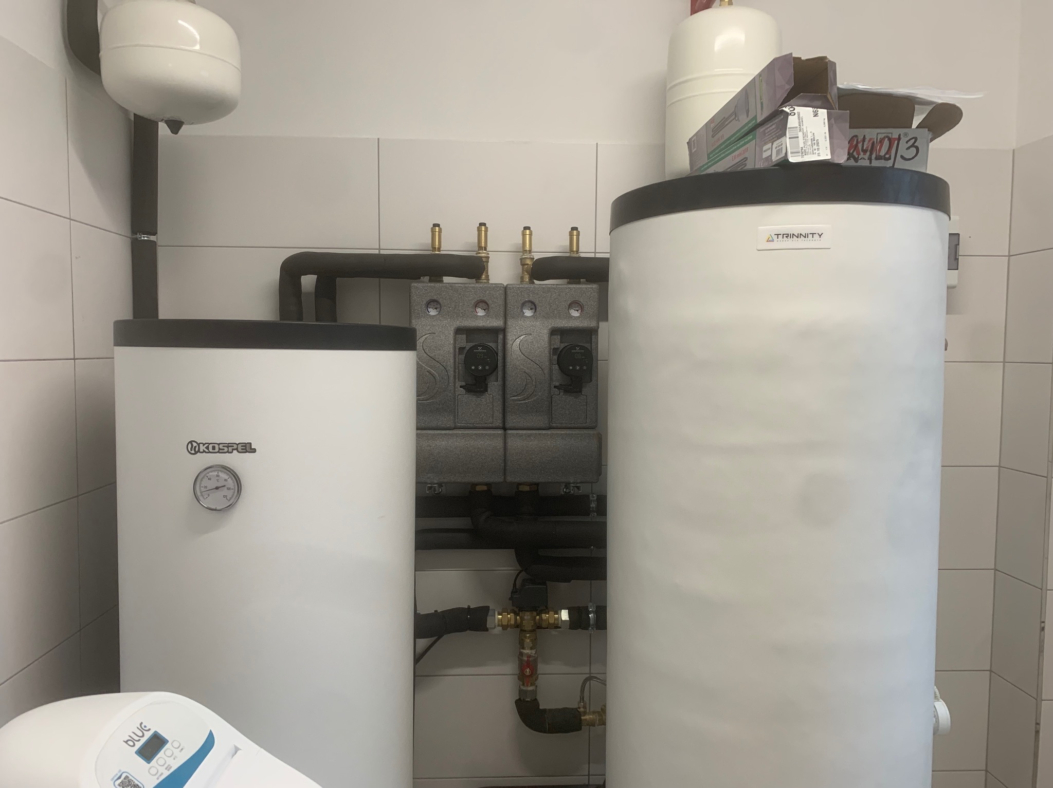 Biały zasobnik ciepłej wody użytkowej Kospel z manometrem i zbiornik Trinity z pudełkami na górze, na tle białych płytek. Widoczne czarne rury i szare zawory.