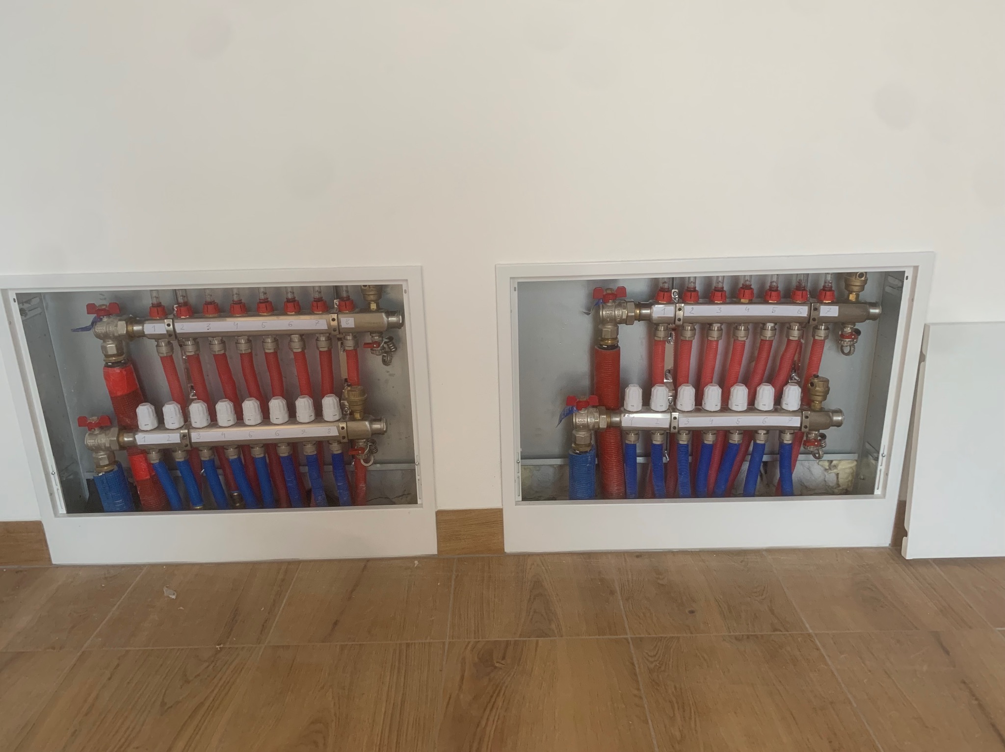 Dwa kolektory ogrzewania podłogowego w białych szafkach, z czerwonymi i niebieskimi rurami, zamontowane na ścianie nad drewnopodobną podłogą.