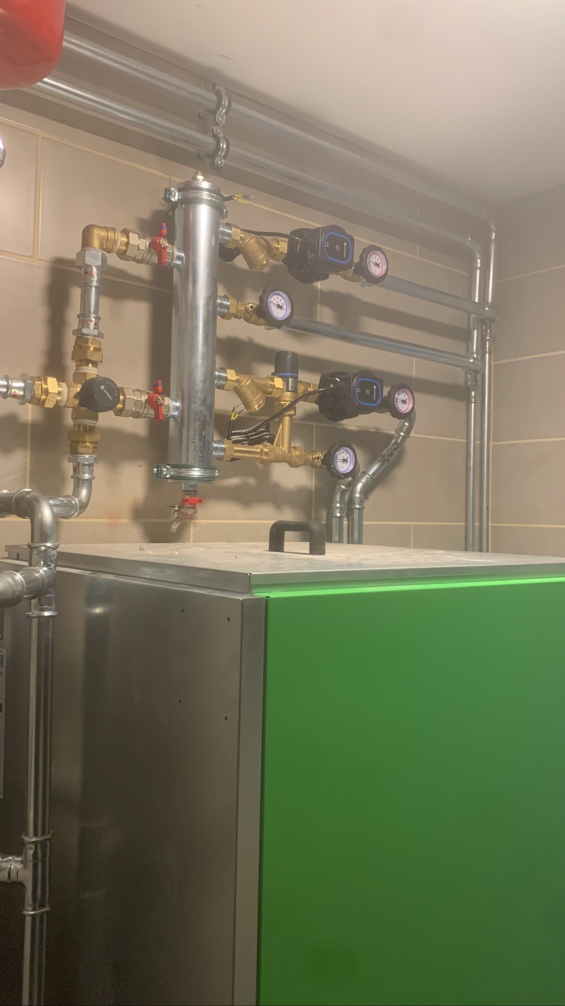 Zielono-srebrny piec z podłączonymi rurami, zaworami i manometrami. Instalacja grzewcza w pomieszczeniu z beżowymi płytkami. Widoczne elementy mocujące rury do sufitu.
