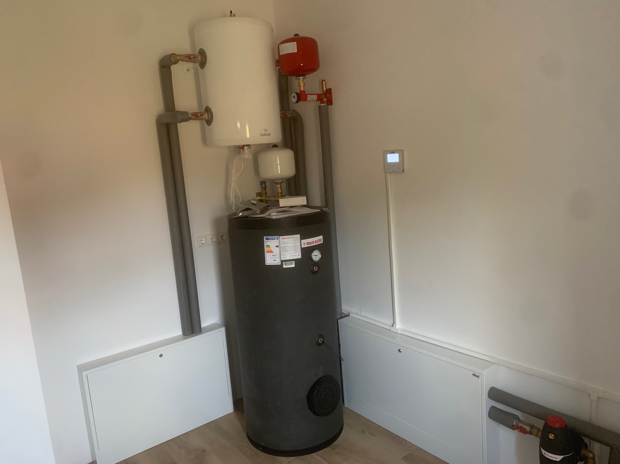 Zestawiony pionowo system grzewczy z czarnym zasobnikiem i białym zbiornikiem wyrównawczym, rury w szarych otulinach, na tle białych ścian i podłogi z paneli.