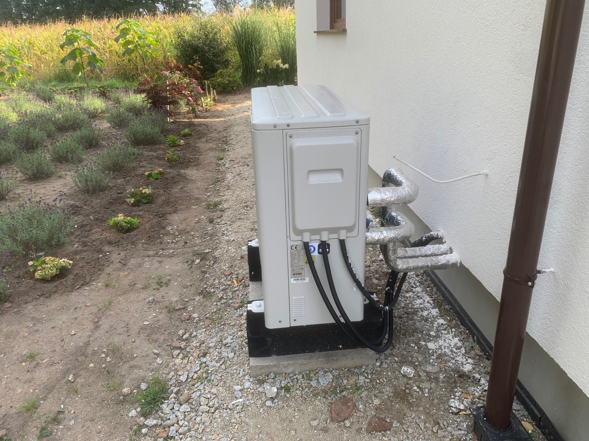 Biała pompa ciepła YORK zamontowana przy ścianie domu, z widocznymi rurami w izolacji. W tle ogród z roślinami i kwiatami. Ujęcie z poziomu gruntu.