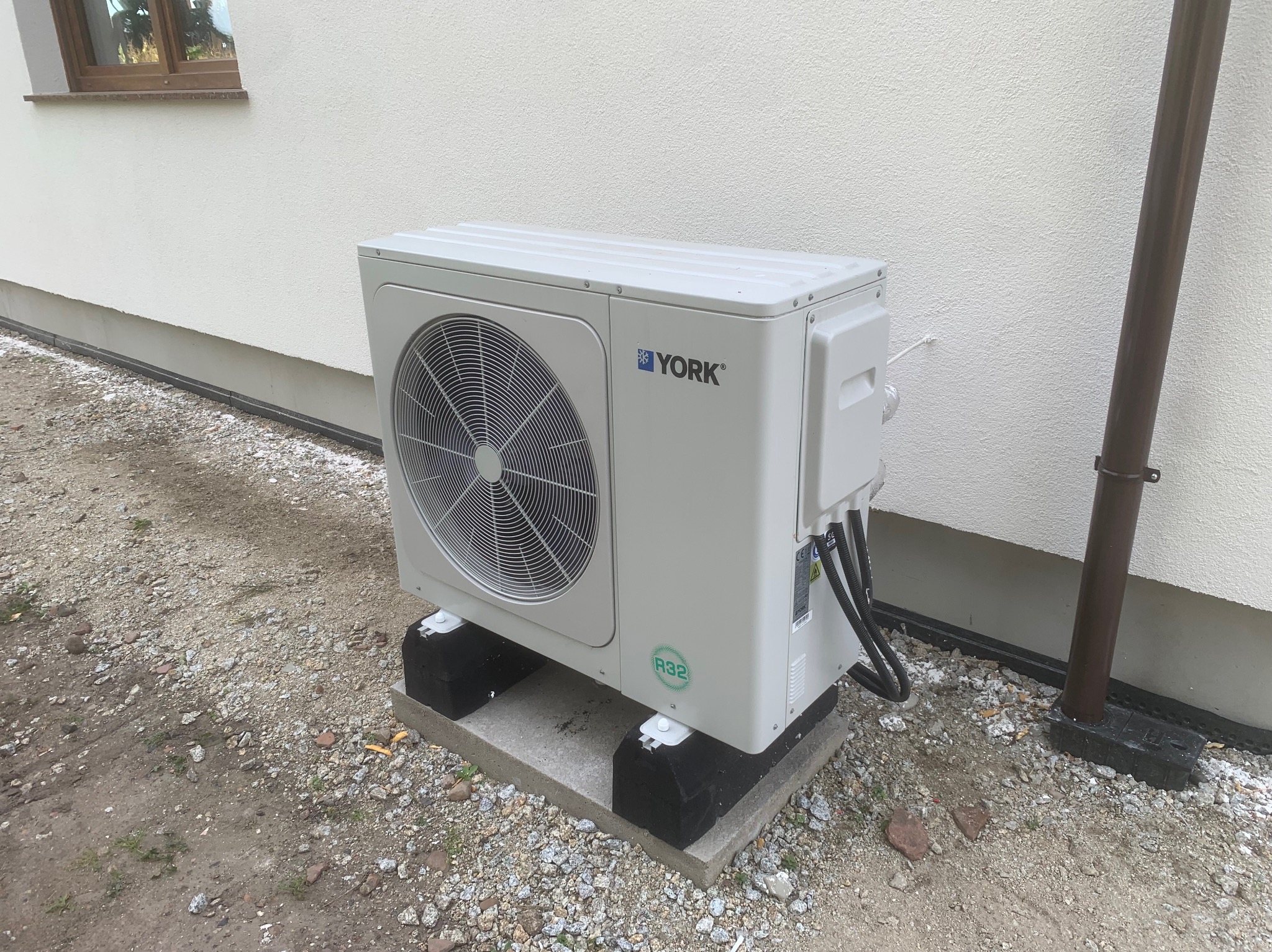 Biała pompa ciepła York R32, zamontowana na betonowej podstawie z czarnymi podkładkami, stoi przy białej ścianie budynku z brązową rurą spustową.