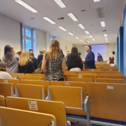 Wykładowca w ciemnej marynarce przemawia do grupy studentów w sali wykładowej z rzędami drewnianych krzeseł z naklejkami 'Tu należy zająć miejsce', rzutnik wyświetla obraz na ekranie w tle.