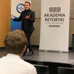 Prelegent w czarnym garniturze gestykuluje na scenie podczas szkolenia z retoryki, za nim baner z napisem 'Akademia Retoryki Igora Zalewskiego' oraz logo PZU, widok zza pleców słuchacza.