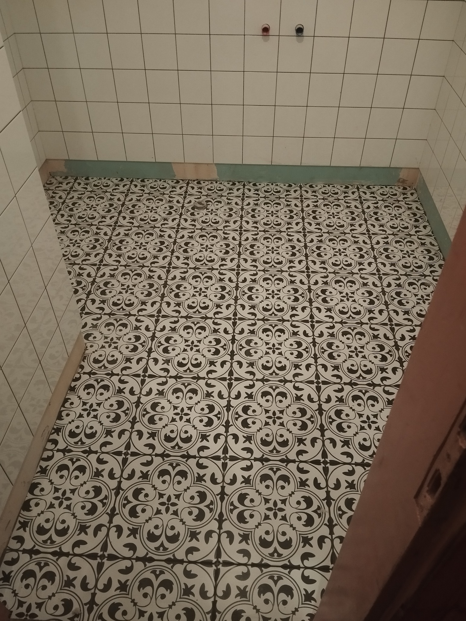 Nowa podłoga z wzorzystych płytek w łazience, z widocznymi fragmentami ściany z białych płytek i przygotowanym podłożem. Widoczne dwa wystające elementy instalacji na ścianie.