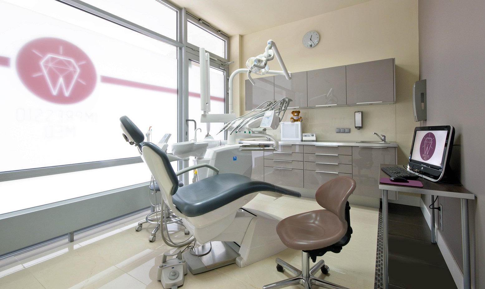 Jasne wnętrze gabinetu stomatologicznego z fotelem dentystycznym, oknem z logo kliniki i szafkami w kolorze taupe, na blacie biurka stoi komputer z widocznym logo kliniki.
