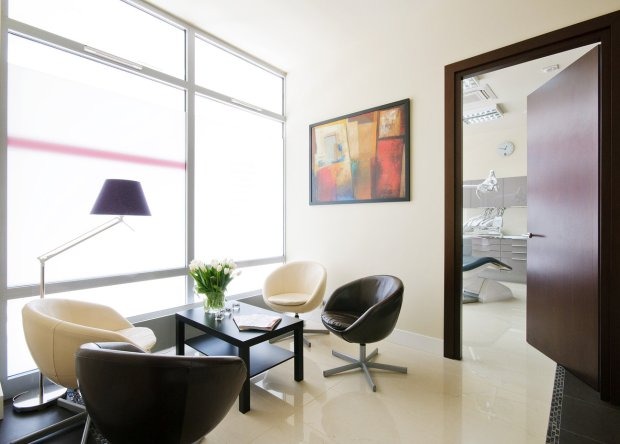 Jasna poczekalnia z czterema krzesłami, stolikiem z tulipanami i widokiem na gabinet przez otwarte drzwi.