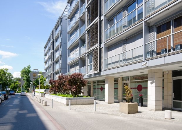 Nowoczesny budynek apartamentowy z balkonami i przeszklonym parterem, widok z ulicy w słoneczny dzień. Na parterze widoczne okna z potencjalnym logo lub grafiką.