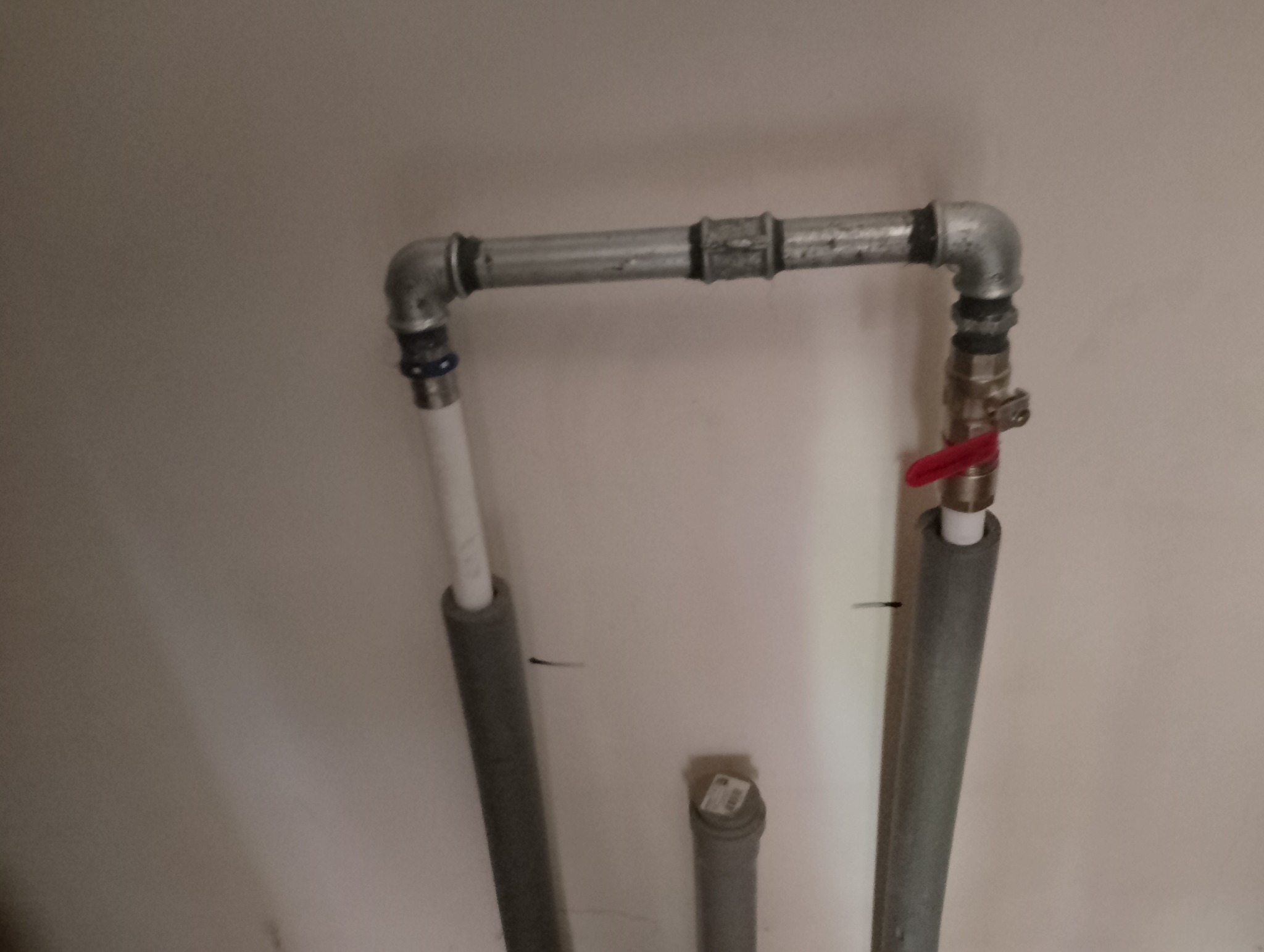 Fragment instalacji hydraulicznej z rurami stalowymi i zaworem z czerwoną rączką, rury pionowe zaizolowane szarą pianką.