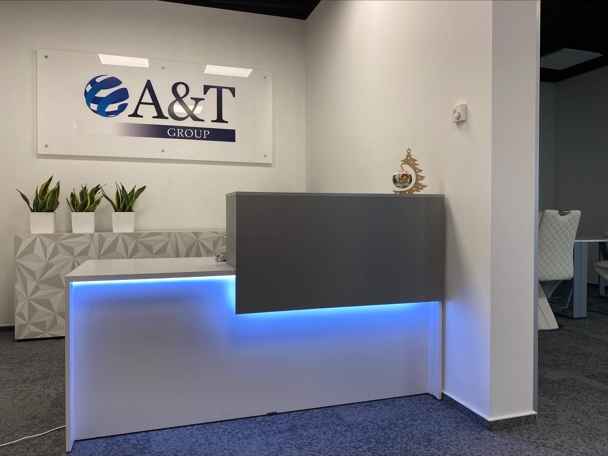 Nowoczesna recepcja biurowa z podświetleniem LED, logo firmy A&T Group na ścianie, dekoracyjne rośliny i ozdoba świąteczna.