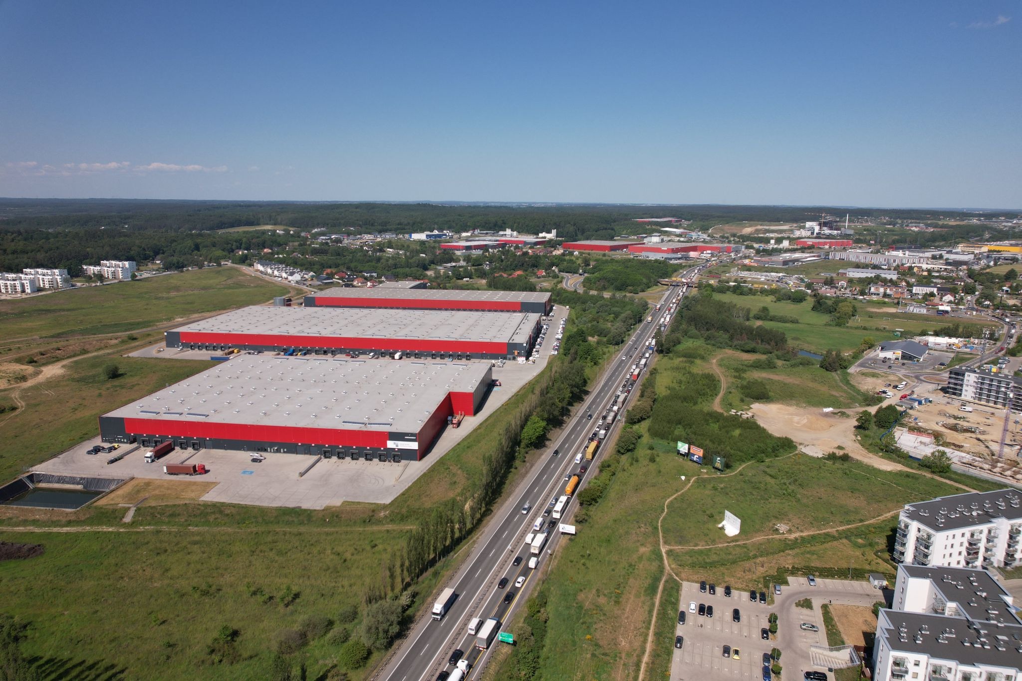 Widok z lotu ptaka na kompleks magazynowy z czerwonymi akcentami, przylegający do zatłoczonej autostrady w słoneczny dzień.
