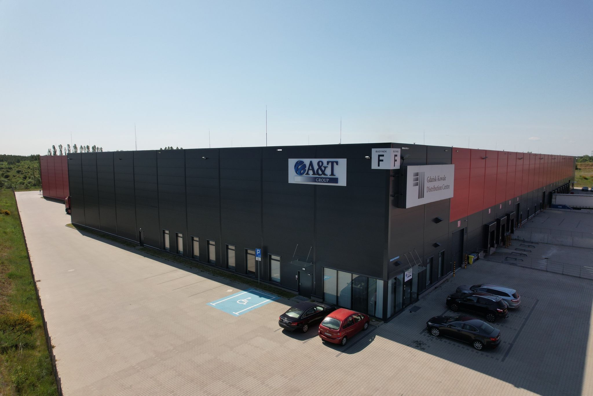 Zewnętrzny widok na nowoczesny budynek magazynowy z czarno-czerwonymi panelami, oznakowany logo 'A&T Group' i tablicą informacyjną 'Gdańsk-Kowale Distribution Centre', z zaparkowanymi samochodami...