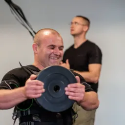 Uśmiechnięty mężczyzna w kamizelce EMS ćwiczy z obciążeniem, w tle drugi mężczyzna obserwuje.