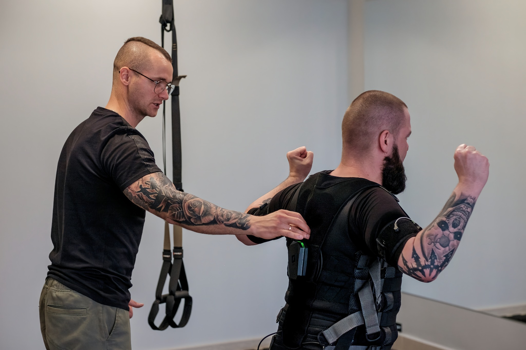 Mężczyzna w okularach z tatuażami na ramieniu dopasowuje elektrostymulację EMS na plecach drugiego mężczyzny z brodą i tatuażami na ramieniu, który stoi z uniesionymi rękami.