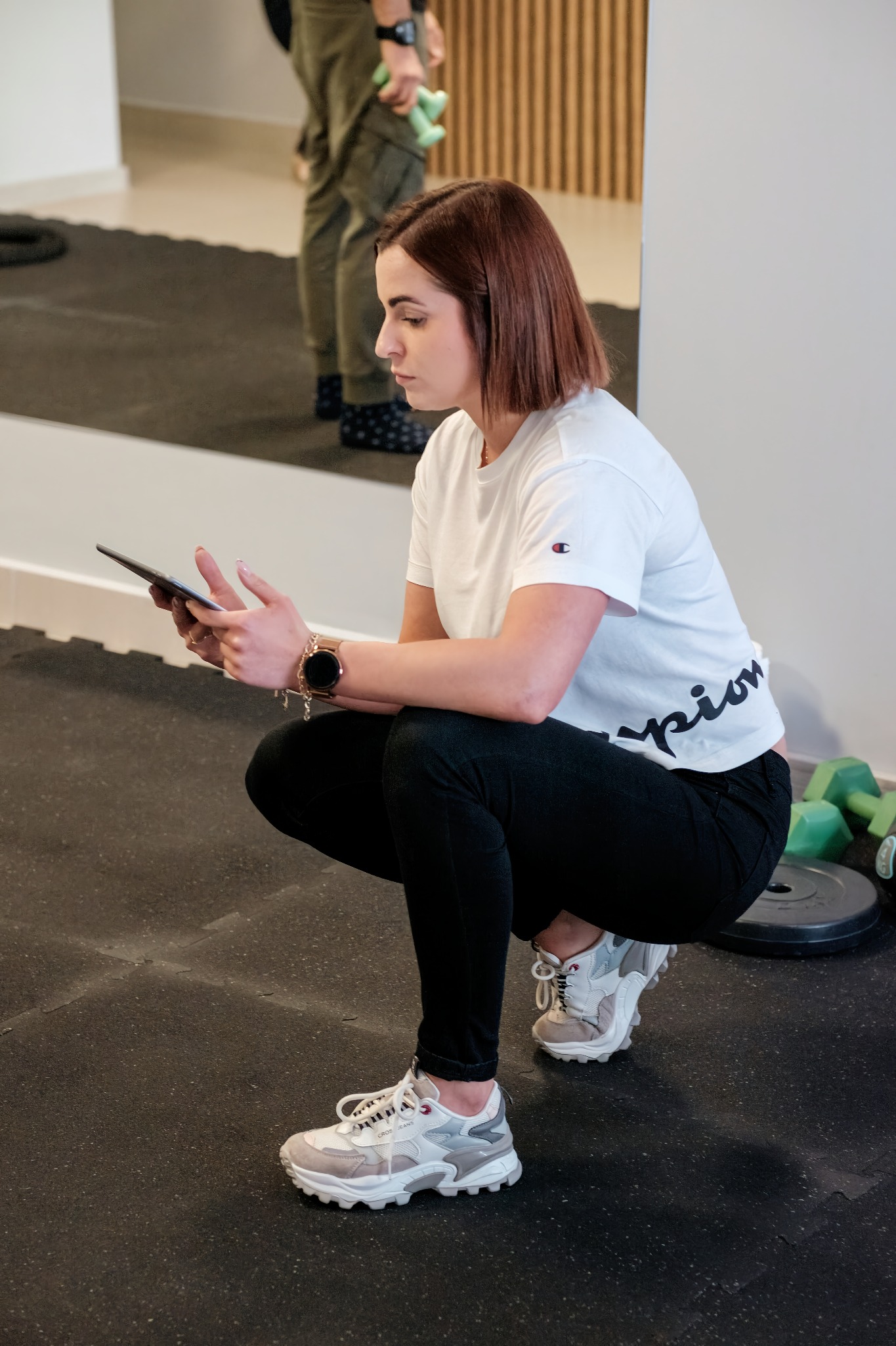 Kobieta w sportowym stroju kuca w siłowni, przeglądając tablet, w tle widoczne ciężarki i osoba z hantlami.