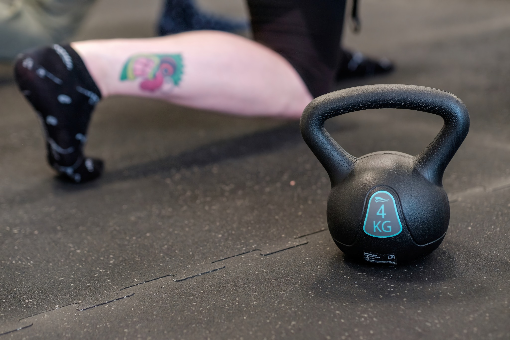 Czarny kettlebell o wadze 4 kg na gumowej podłodze, w tle noga osoby ćwiczącej z widocznym tatuażem na łydce.