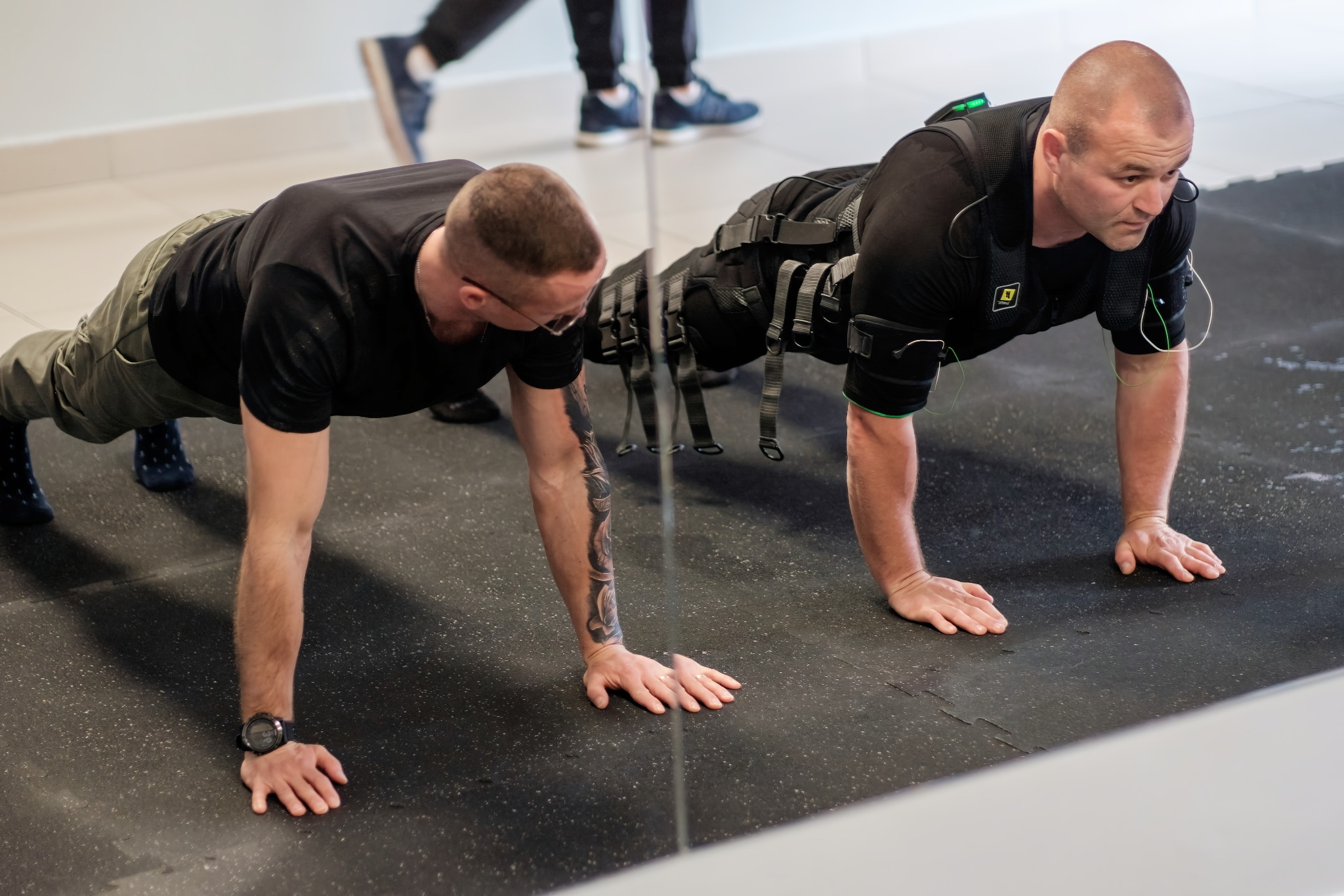 Dwaj mężczyźni wykonują pompki w siłowni. Jeden z nich ma na sobie kamizelkę EMS z elektrodami, drugi bez. Widoczne odbicie w lustrze.