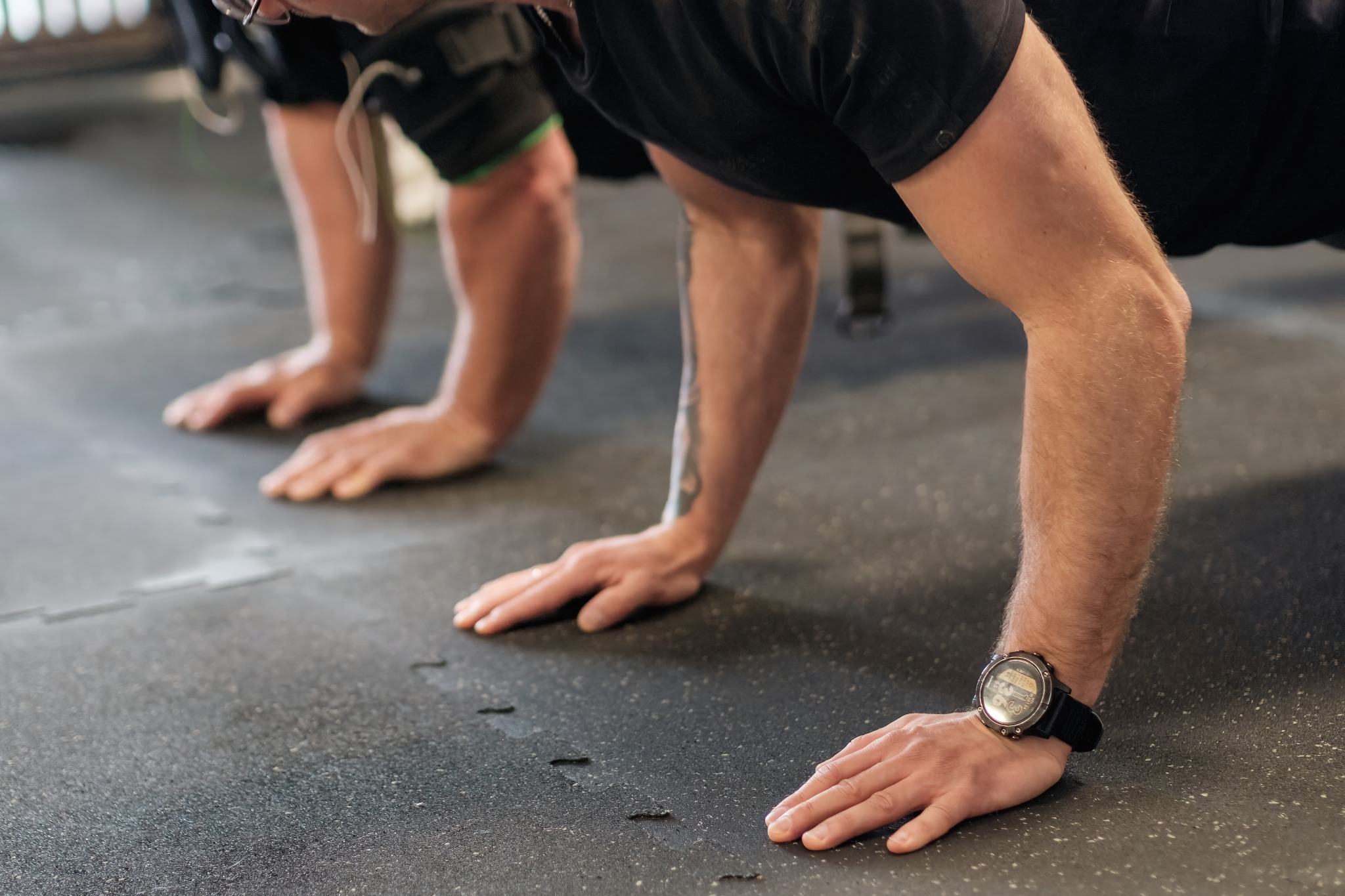 Dwie osoby wykonują pompki na gumowej podłodze w siłowni, z bliska widoczne ręce i przedramiona, na nadgarstku zegarek sportowy.