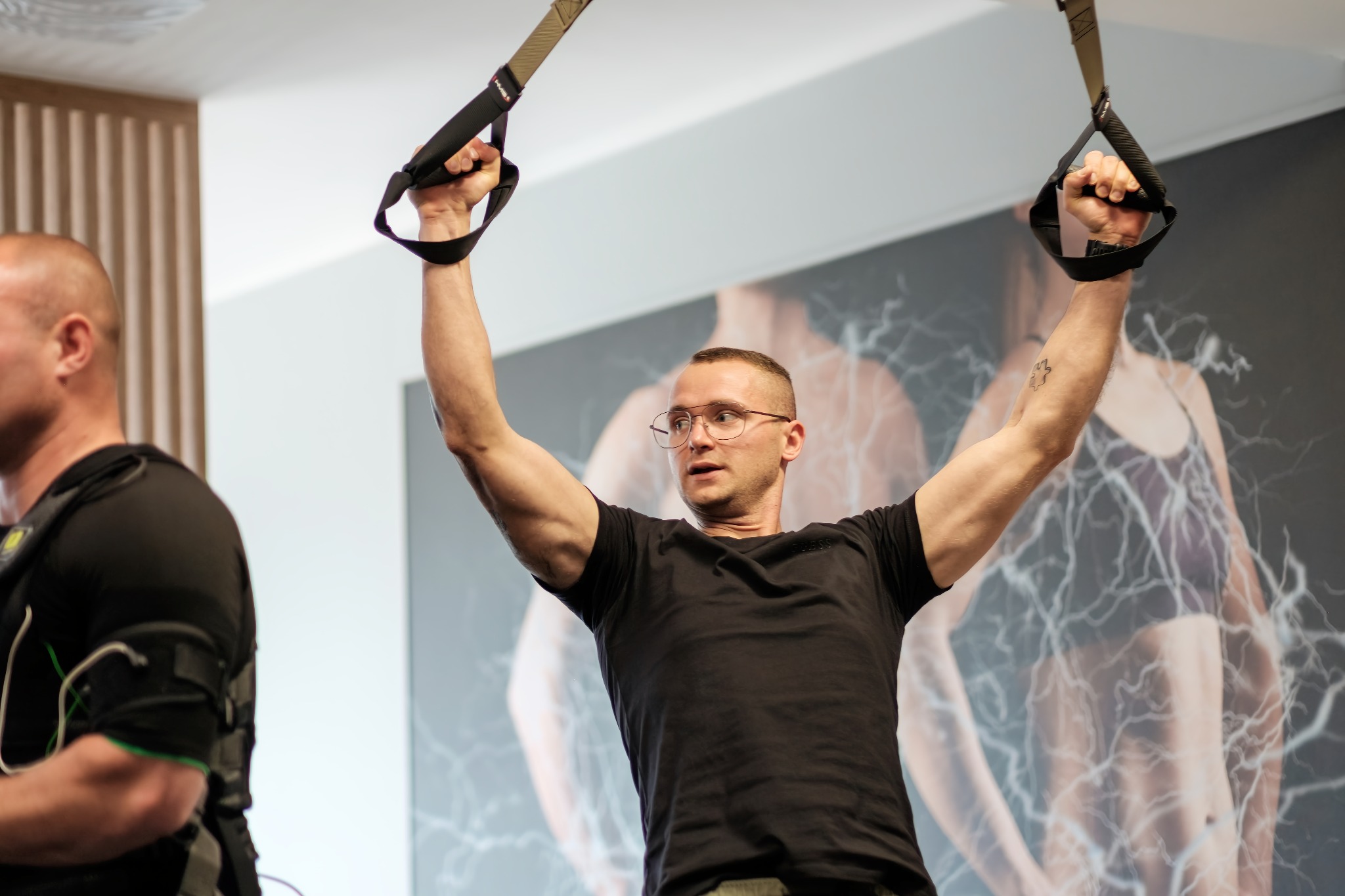Mężczyzna w okularach wykonuje ćwiczenie na taśmach TRX, w tle widoczny mężczyzna z elektrostymulacją mięśni.
