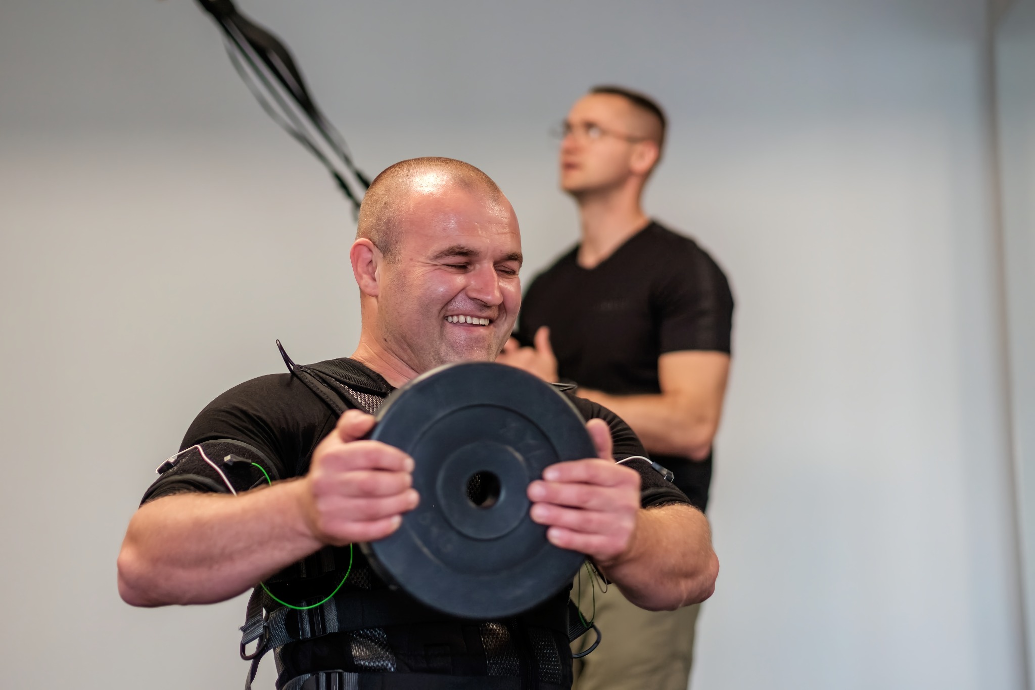 Uśmiechnięty mężczyzna w kamizelce EMS ćwiczy z obciążeniem, w tle drugi mężczyzna obserwuje.