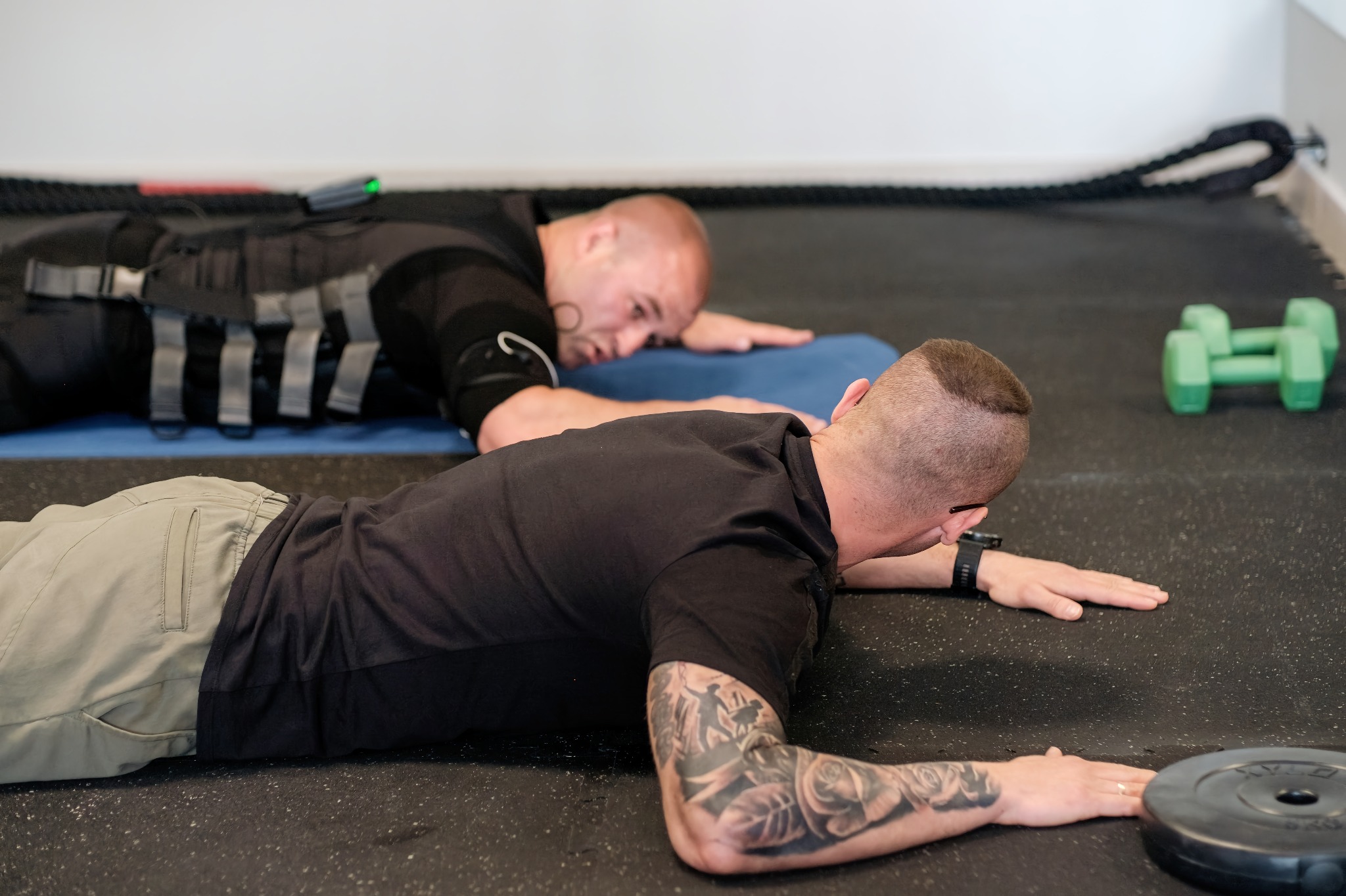 Dwaj mężczyźni leżą na brzuchu na podłodze w siłowni podczas treningu EMS, jeden z widocznym tatuażem na ramieniu, obok leżą zielone hantle i lina do ćwiczeń.