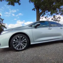 Srebrny sedan Toyota Corolla na żwirowym podłożu z widocznym błękitnym niebem i drzewami w tle.