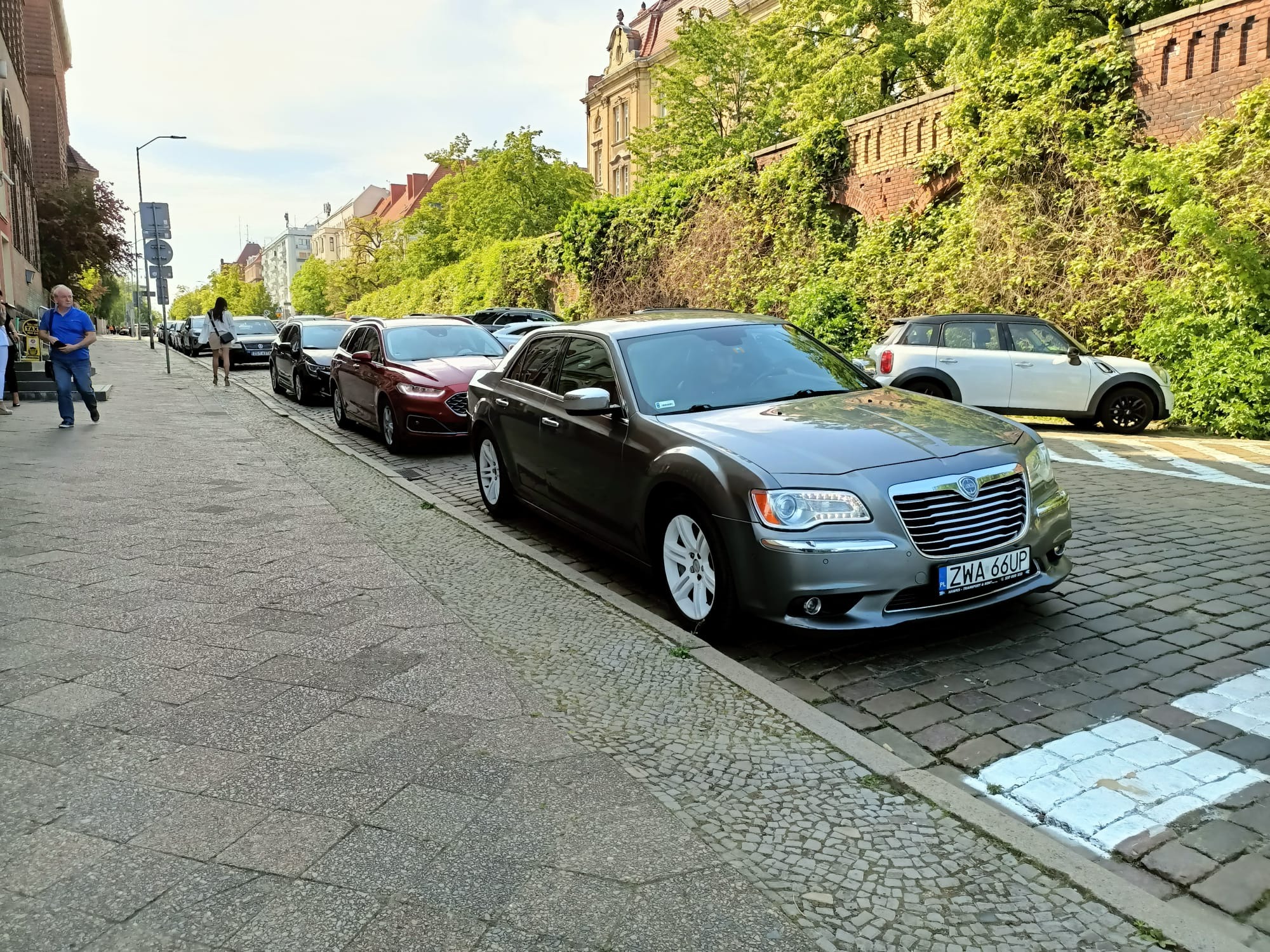 Szary samochód Chrysler 300 parkuje na brukowanej ulicy w mieście, widoczne białe felgi i tablica rejestracyjna ZWA 66UP, w tle budynki i zieleń.