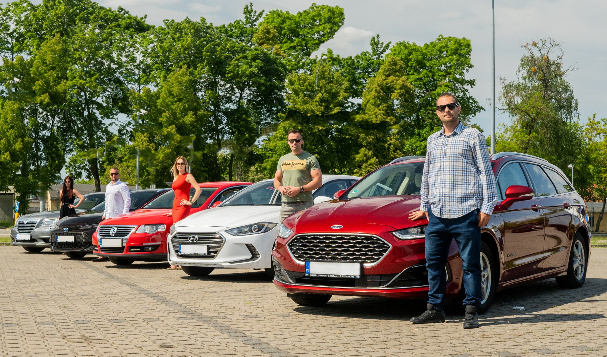 Flota samochodów osobowych różnych marek, z pozującymi osobami, na tle zieleni drzew.