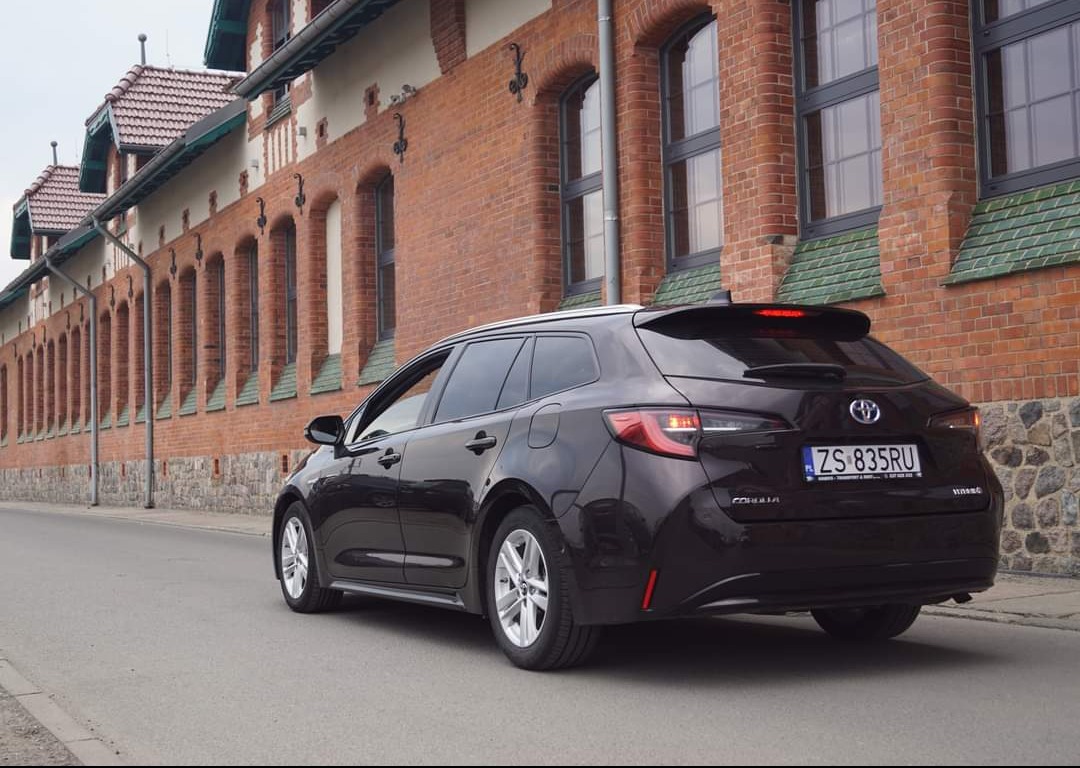 Ciemnobrązowa Toyota Corolla kombi zaparkowana na ulicy przed budynkiem z czerwonej cegły z rzędem łukowych okien.