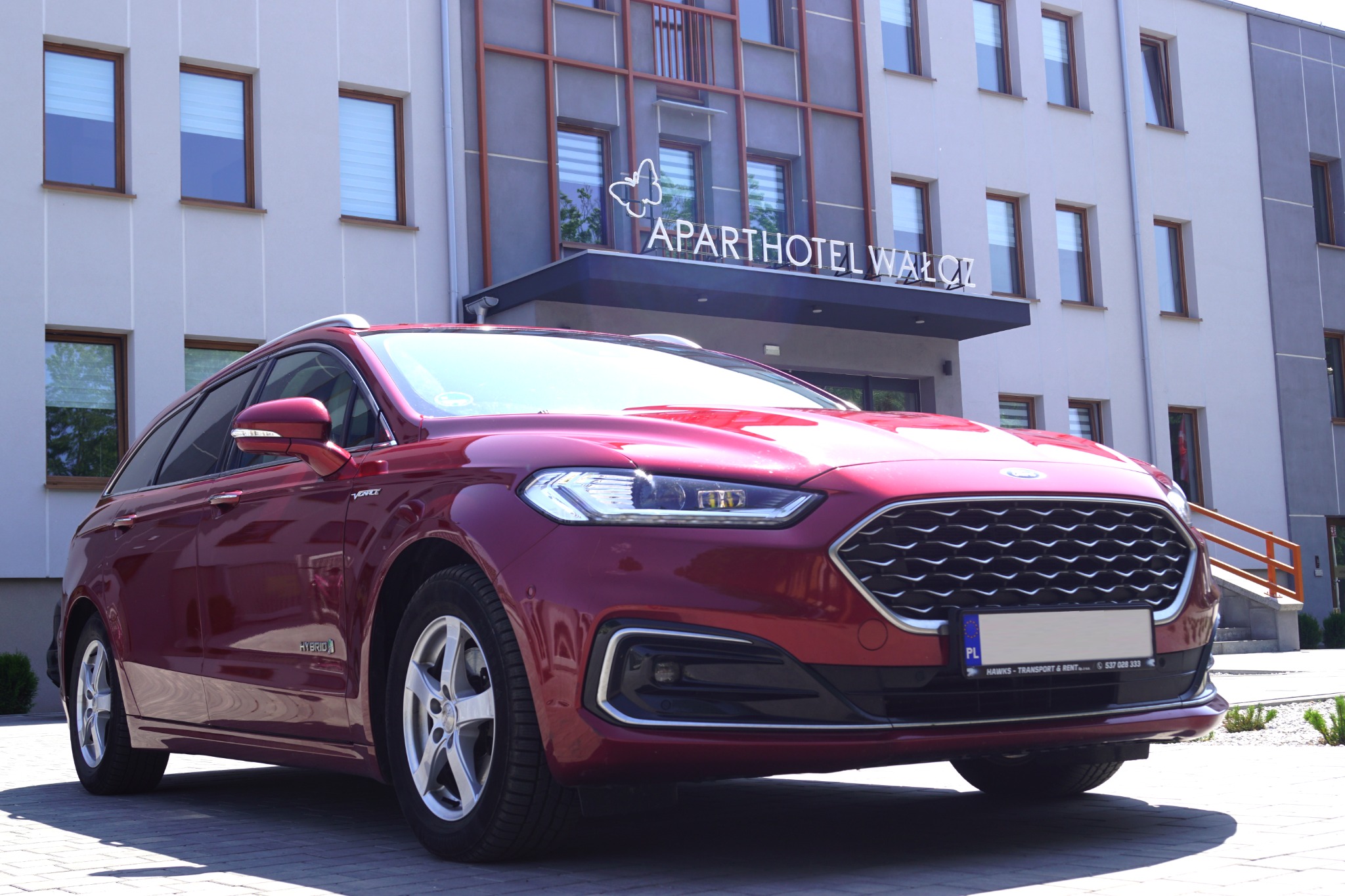 Czerwony samochód typu kombi z napędem hybrydowym zaparkowany przed budynkiem Aparthotelu Wałcz w słoneczny dzień.